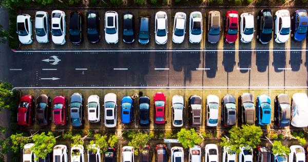 Aerial view of a mostly full parking lot with rows of parked cars, directional arrows on the ground, and trees interspersed between some rows