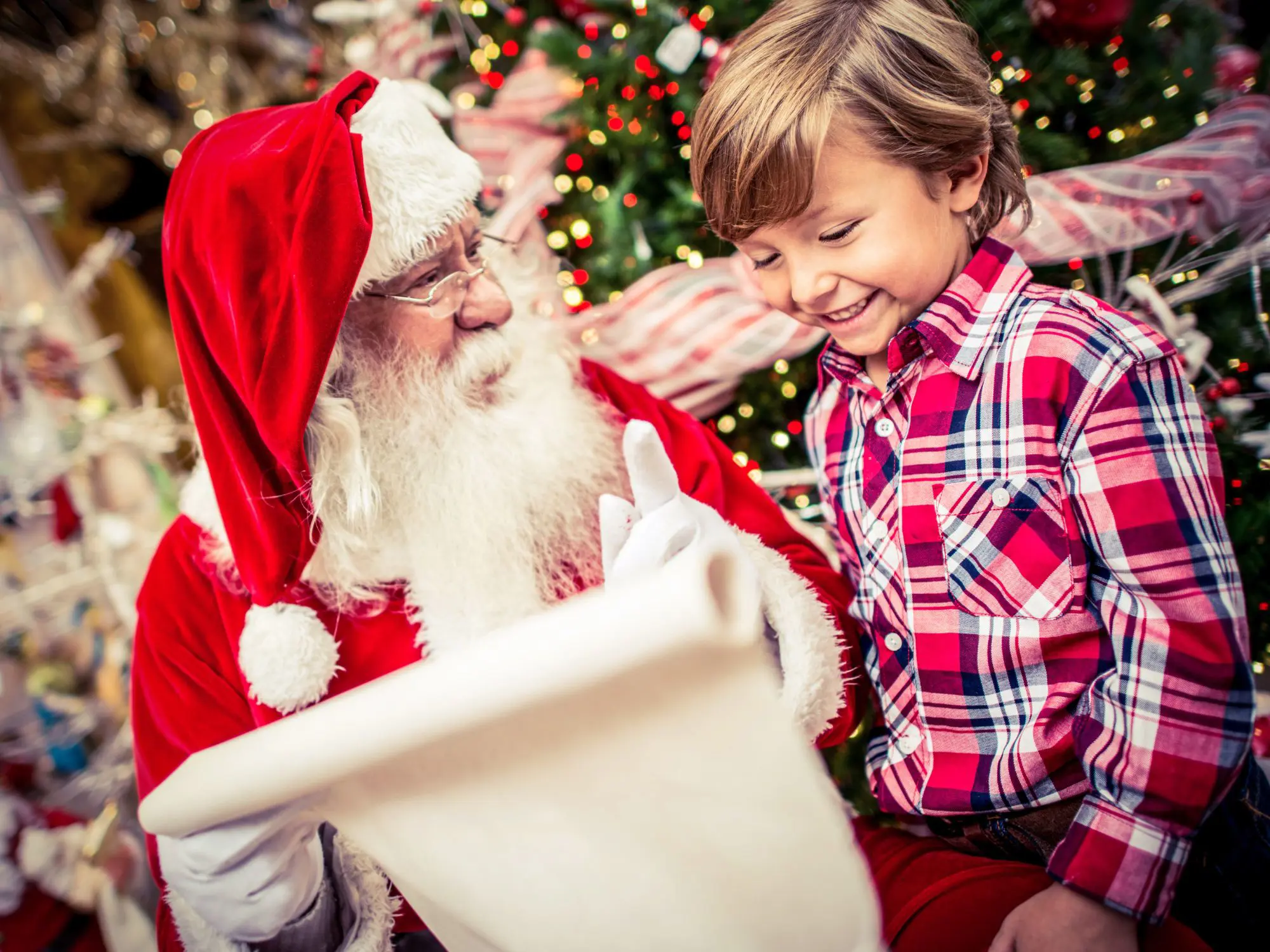 A person dressed as Santa Claus holding a large scroll while sitting next to a child wearing a red and white plaid shirt. Behind them is a decorated Christmas tree with ornaments and lights.