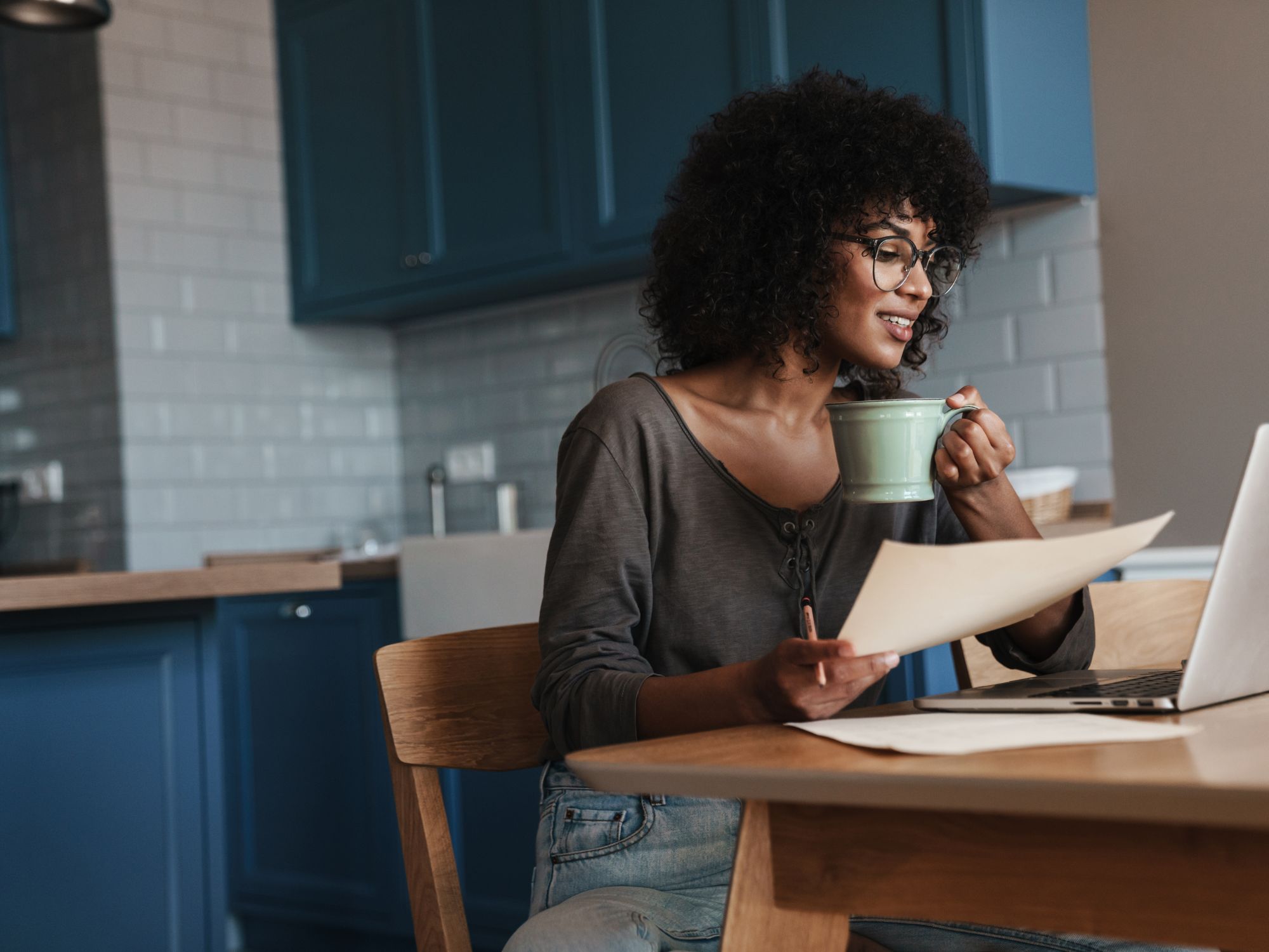 Person sitting at a wooden kitchen table with blue cabinets and white tiled backsplash, holding a green mug and a piece of paper while looking at an open laptop. Wearing glasses, a grey top, and light blue jeans, suggesting a relaxed work-from-home or study setting.