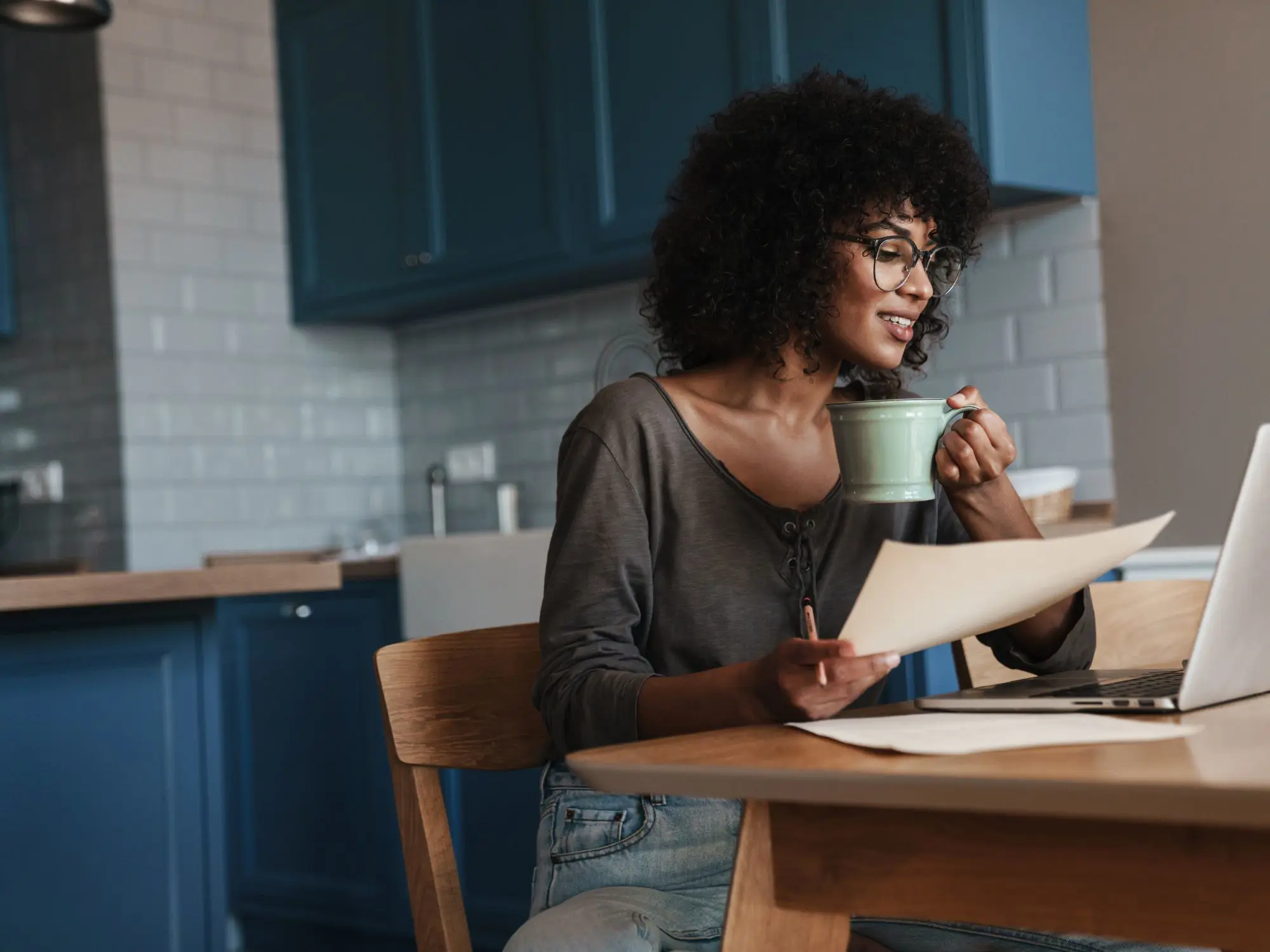 Person sitting at a wooden kitchen table with blue cabinets and white tiled backsplash, holding a green mug and a piece of paper while looking at an open laptop. Wearing glasses, a grey top, and light blue jeans, suggesting a relaxed work-from-home or study setting.