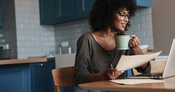 Person sitting at a wooden kitchen table with blue cabinets and white tiled backsplash, holding a green mug and a piece of paper while looking at an open laptop. Wearing glasses, a grey top, and light blue jeans, suggesting a relaxed work-from-home or study setting.