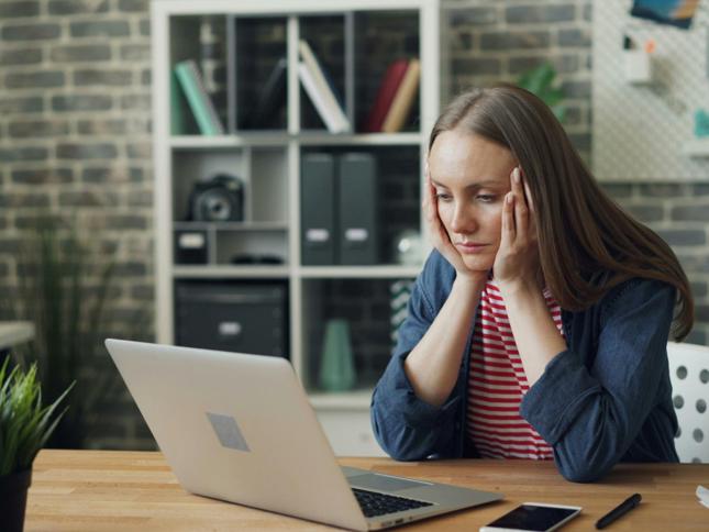 A person sitting at a desk with a laptop open in front, hands resting on the sides of the head. A smartphone and pen are on the desk, and shelves with books and storage boxes are in the background. A person sitting at a desk with a laptop open in front, hands resting on the sides of the head. A smartphone and pen are on the desk, and shelves with books and storage boxes are in the background.