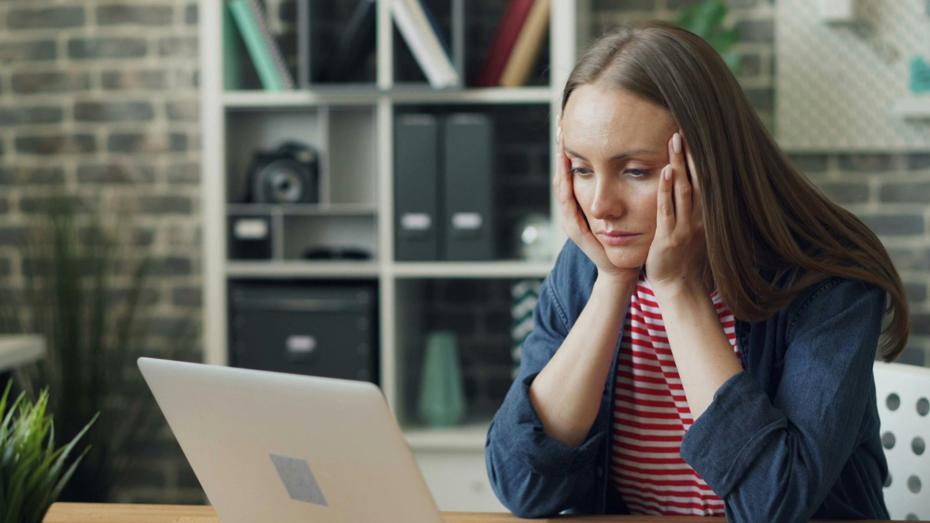A person sitting at a desk with a laptop open in front, hands resting on the sides of the head. A smartphone and pen are on the desk, and shelves with books and storage boxes are in the background. A person sitting at a desk with a laptop open in front, hands resting on the sides of the head. A smartphone and pen are on the desk, and shelves with books and storage boxes are in the background.