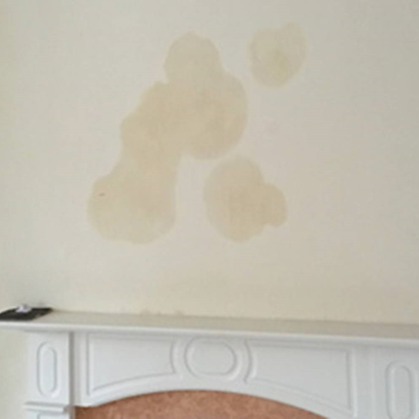 Wall above a white fireplace mantel showing large, irregular damp stains, suggesting water seepage or leakage.