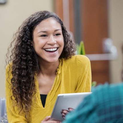 Two people sitting indoors and conversing. One wears a yellow top and holds a tablet; the other wears a green plaid shirt. A door and plants are visible in the blurred background.