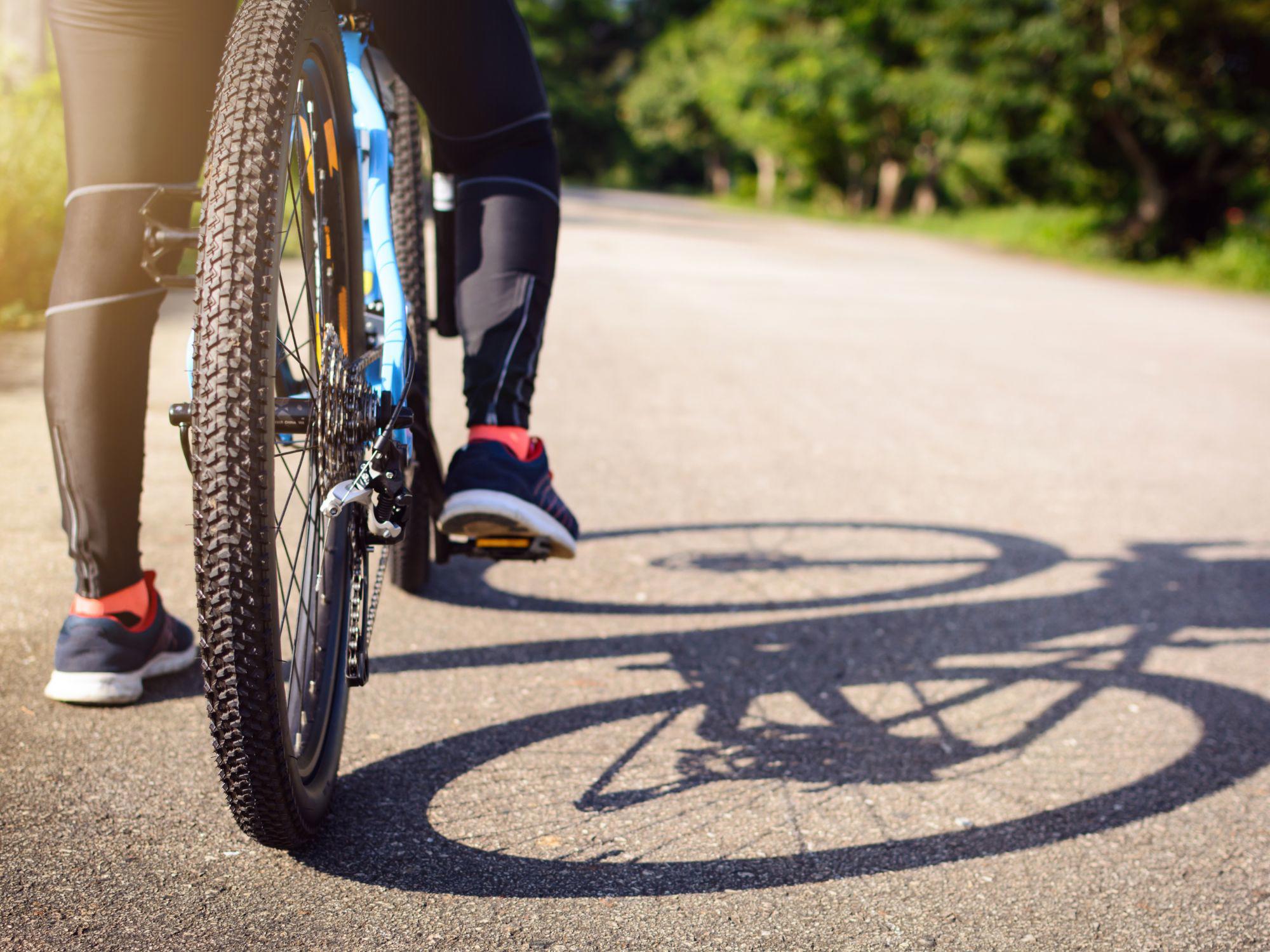 A close-up of a cyclist’s legs and bike on a sunlit paved road. The rider wears tight cycling trousers and athletic shoes. The bike has thick, knobby tires, and shadows stretch across the road. Trees and greenery line the route in the background.