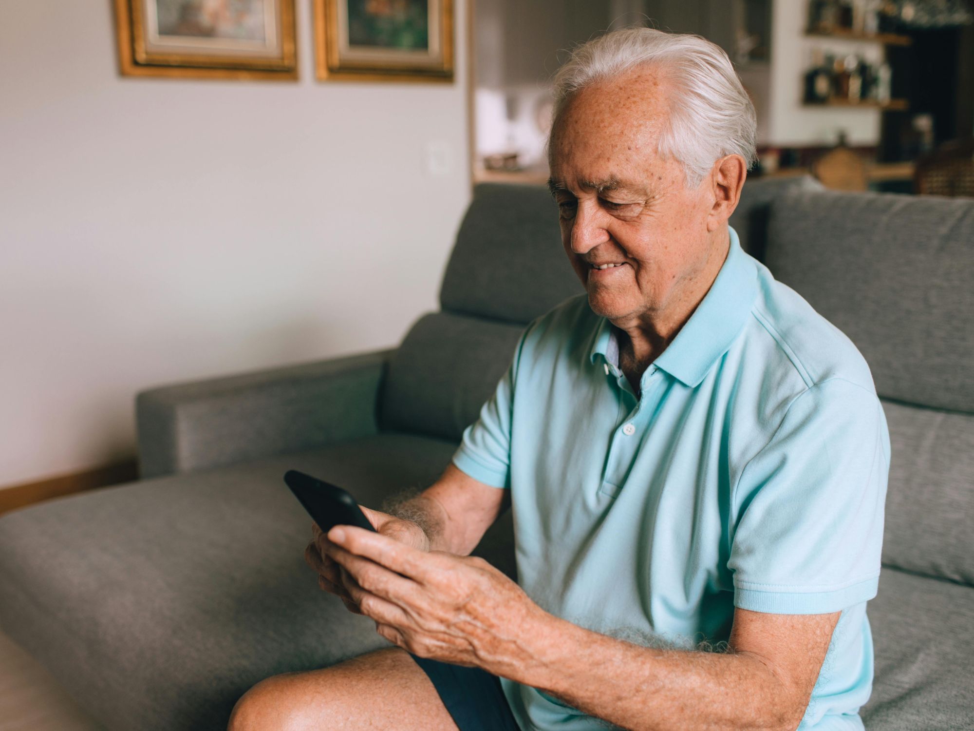An elderly person in a light blue polo shirt and dark shorts sits on a couch, looking at a smartphone. Behind them are two framed pictures on the wall and shelves with various household items.