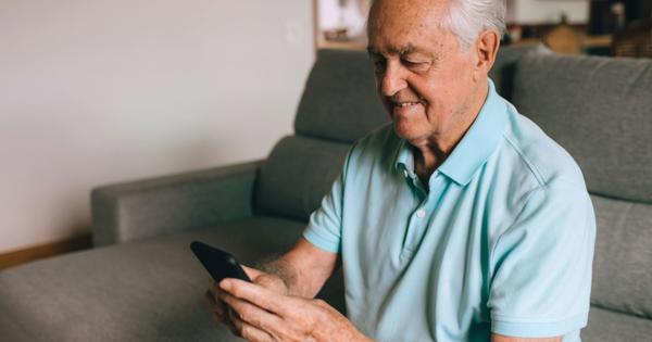 An elderly person in a light blue polo shirt and dark shorts sits on a couch, looking at a smartphone. Behind them are two framed pictures on the wall and shelves with various household items.