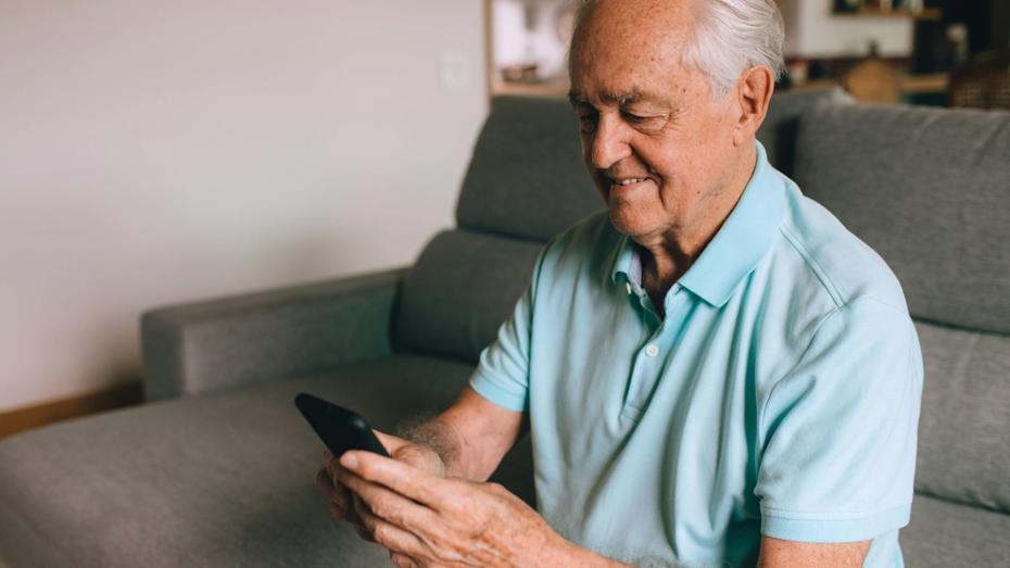 An elderly person in a light blue polo shirt and dark shorts sits on a couch, looking at a smartphone. Behind them are two framed pictures on the wall and shelves with various household items. An elderly person in a light blue polo shirt and dark shorts sits on a couch, looking at a smartphone. Behind them are two framed pictures on the wall and shelves with various household items.