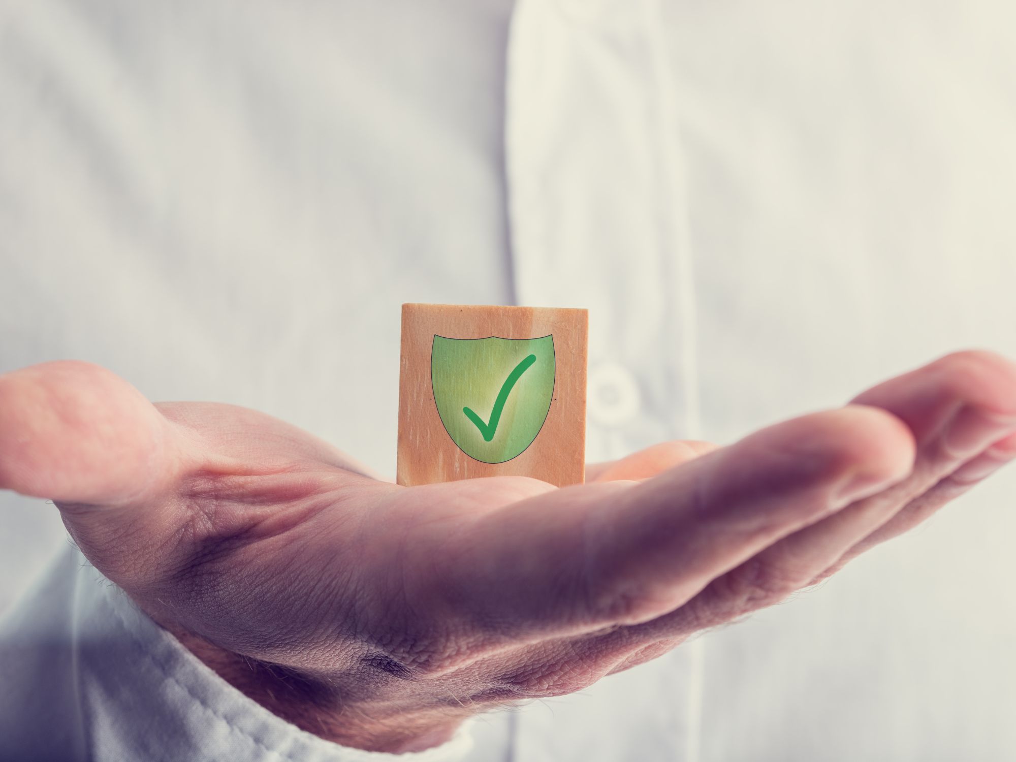 Open hand holding a wooden block with a green shield and checkmark, symbolising protection, trust, and safeguarding within the community.