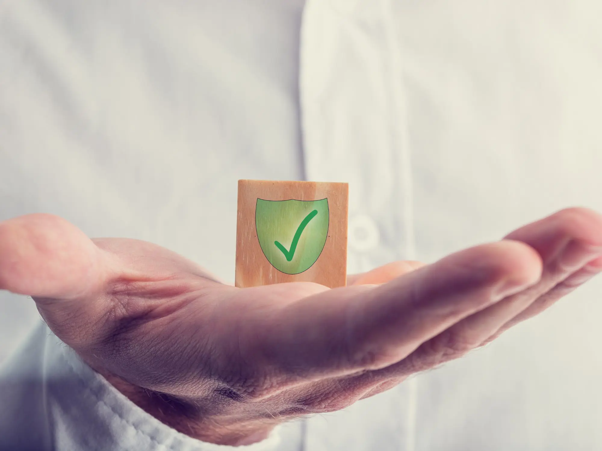 Open hand holding a wooden block with a green shield and checkmark, symbolising protection, trust, and safeguarding within the community.