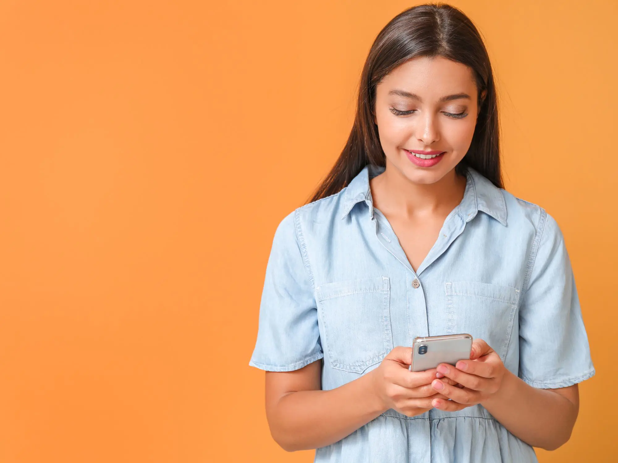 Person standing against an orange background, wearing a light blue short-sleeved button-up shirt, looking down at a smartphone held in both hands, appearing focused on the screen.