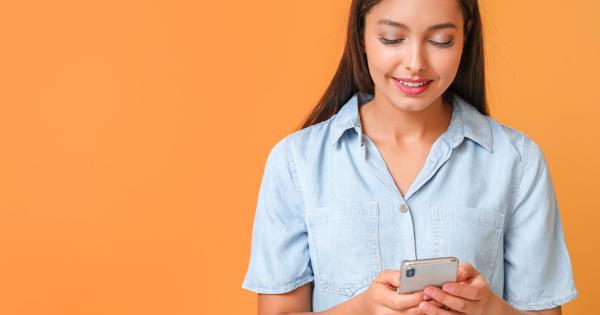 Person standing against an orange background, wearing a light blue short-sleeved button-up shirt, looking down at a smartphone held in both hands, appearing focused on the screen.