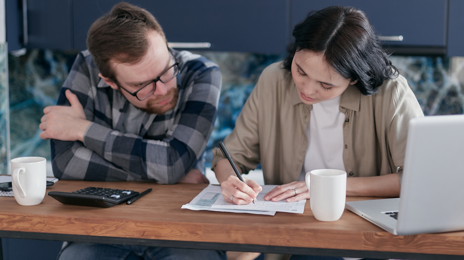 Two people sitting at a table reviewing paperwork together, with one person writing on a form. A laptop, calculator and mugs are on the table.