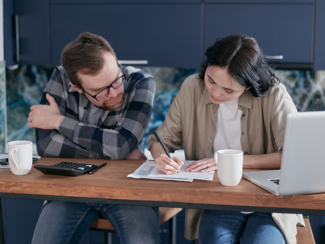 Two people sitting at a table reviewing paperwork together, with one person writing on a form. A laptop, calculator and mugs are on the table.