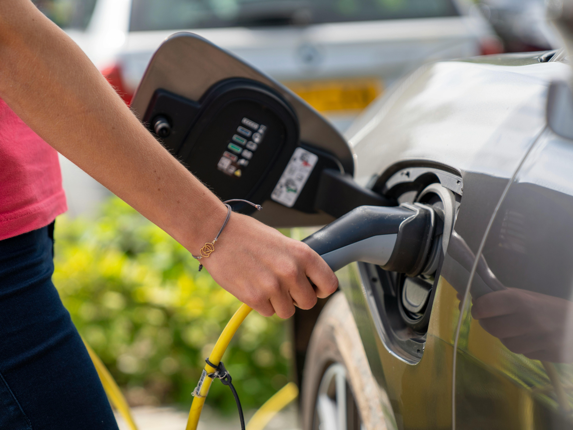 Close-up of a hand holding an electric vehicle charging plug connected to a car, with greenery blurred in the background.
