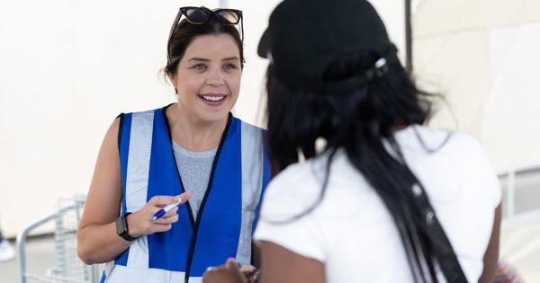 Two people interacting outdoors. One wears a blue and white safety vest with sunglasses on their head; the other wears a black cap and white shirt. Equipment is visible in the background.
