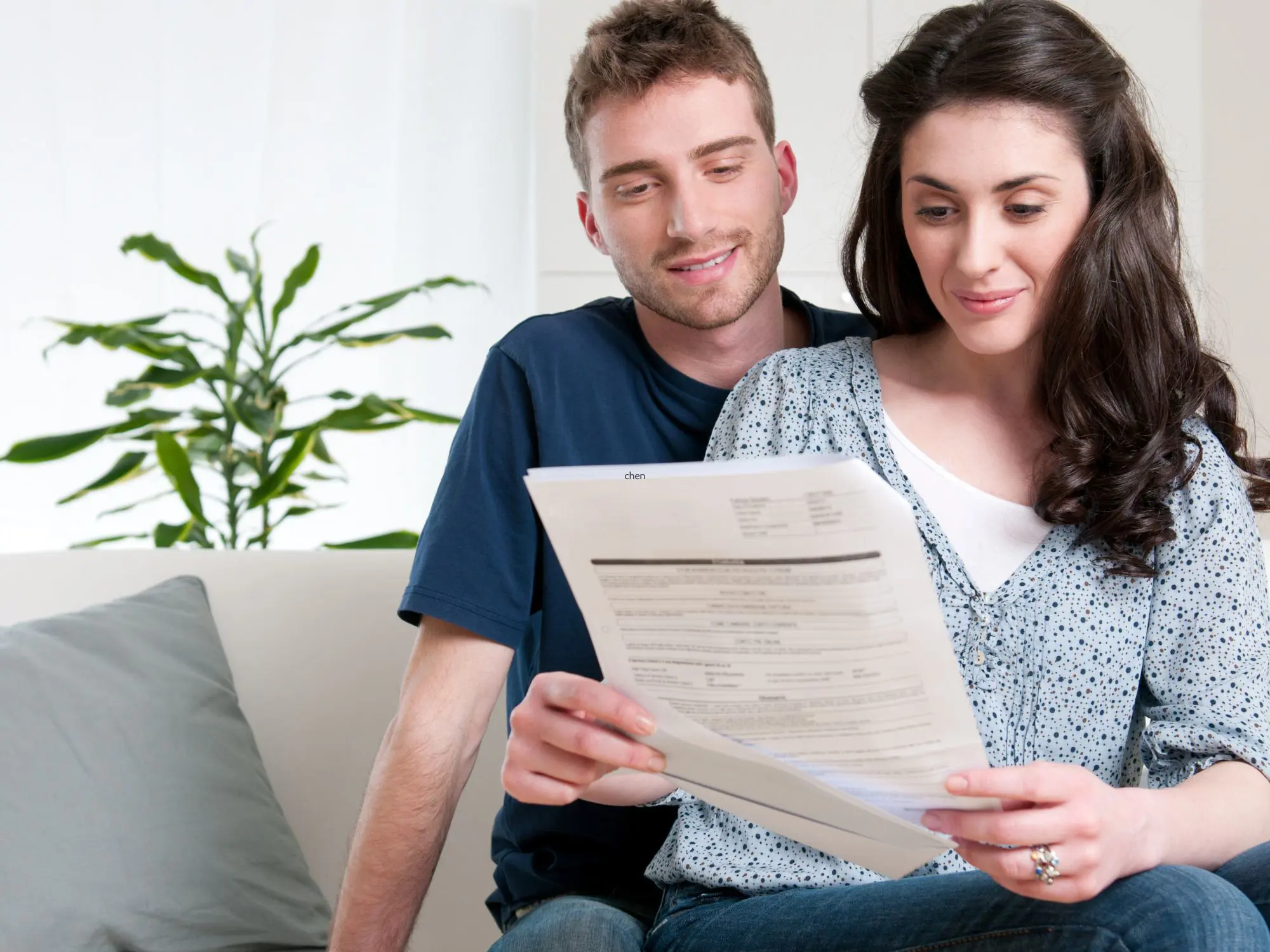 Two people sitting on a couch reviewing a document together, with a plant in the background and a grey cushion nearby.