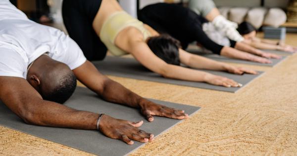 Group of people in child's pose on yoga mats in a studio with wooden floors and shelves holding towels and yoga equipment.
