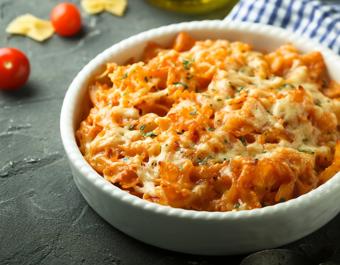 A white ceramic bowl filled with cheesy baked pasta in a tomato sauce, garnished with herbs. The bowl is placed on a dark surface with cherry tomatoes, uncooked pasta, and a blue and white checkered cloth nearby.