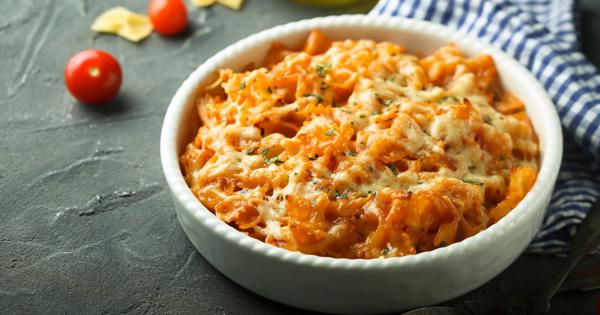 A white ceramic bowl filled with cheesy baked pasta in a tomato sauce, garnished with herbs. The bowl is placed on a dark surface with cherry tomatoes, uncooked pasta, and a blue and white checkered cloth nearby.