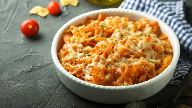 A white ceramic bowl filled with cheesy baked pasta in a tomato sauce, garnished with herbs. The bowl is placed on a dark surface with cherry tomatoes, uncooked pasta, and a blue and white checkered cloth nearby.