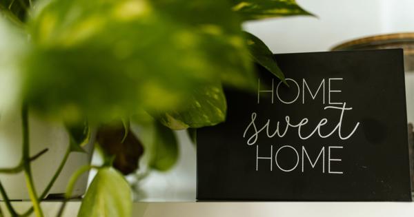 A black sign with white text reading 'HOME SWEET HOME' placed on a white shelf, partially obscured by green plant leaves on the left side.