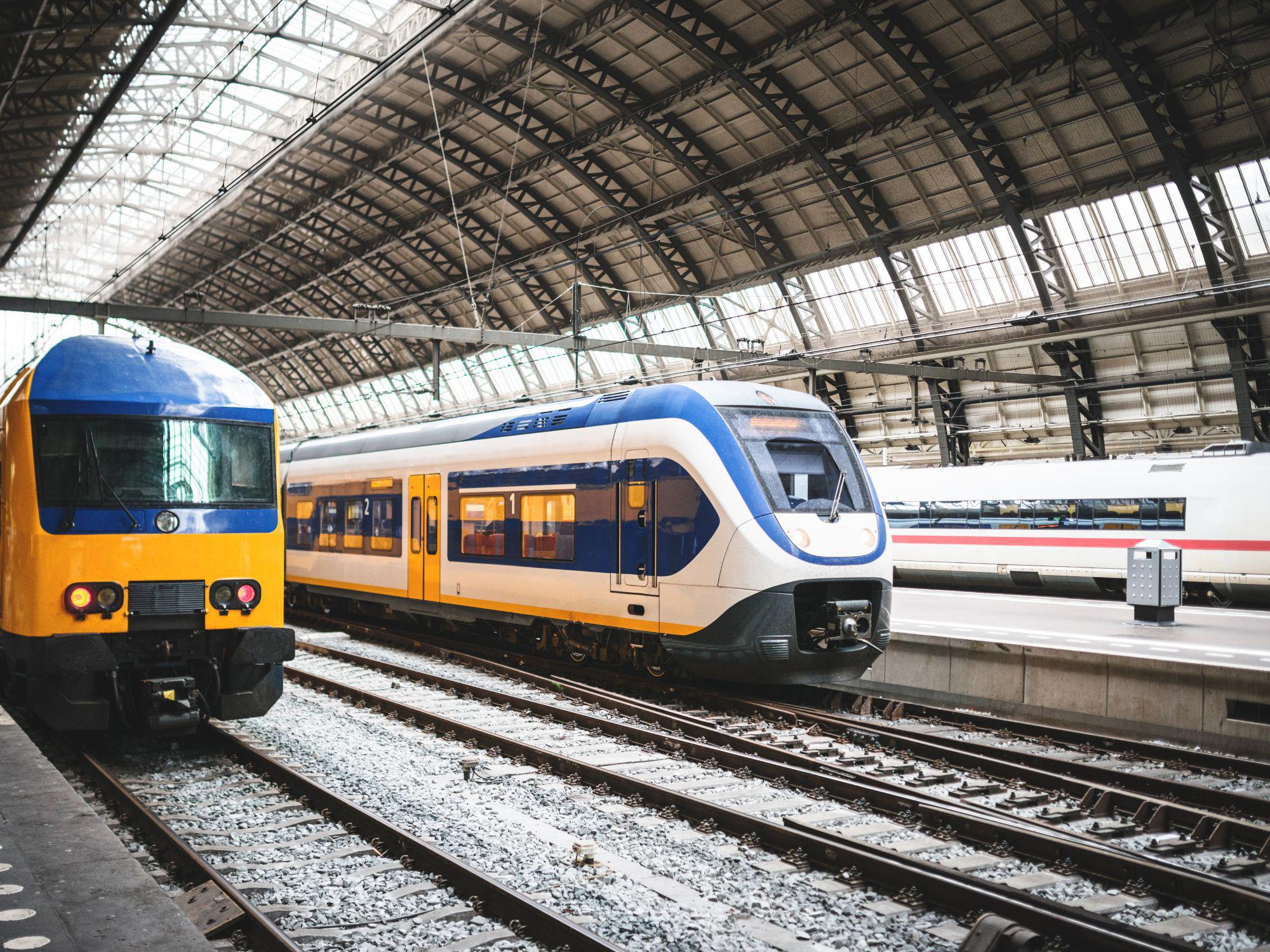 Three trains at a modern station beneath a large arched glass roof. The left train is yellow and blue; the centre train is white with blue and yellow accents; and a third train in the background is white with red stripes.