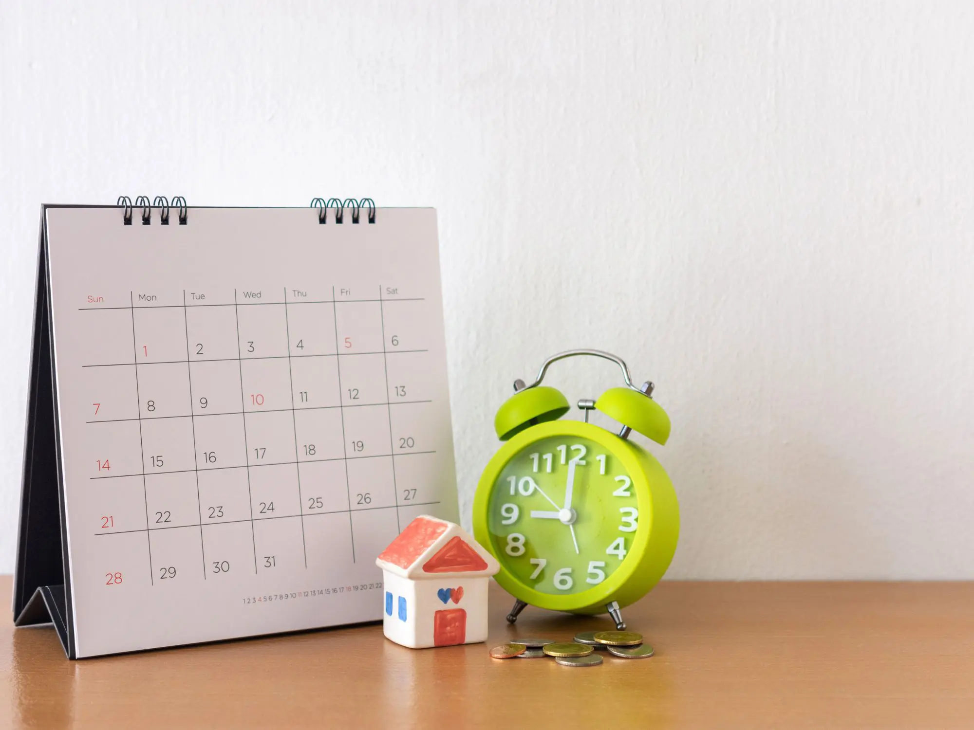 A desk with a February calendar, a green alarm clock showing 10:10, a small model house with a red roof and blue windows, and several coins placed nearby.
