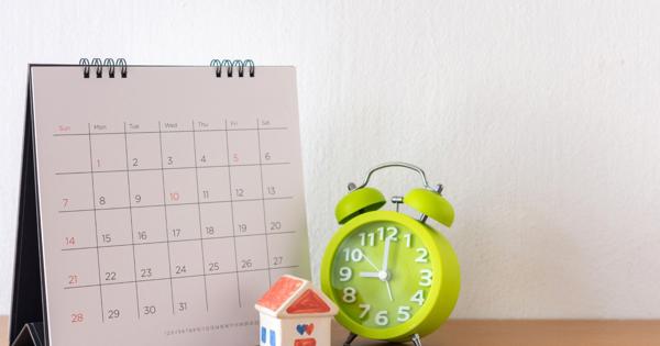 A desk with a February calendar, a green alarm clock showing 10:10, a small model house with a red roof and blue windows, and several coins placed nearby.