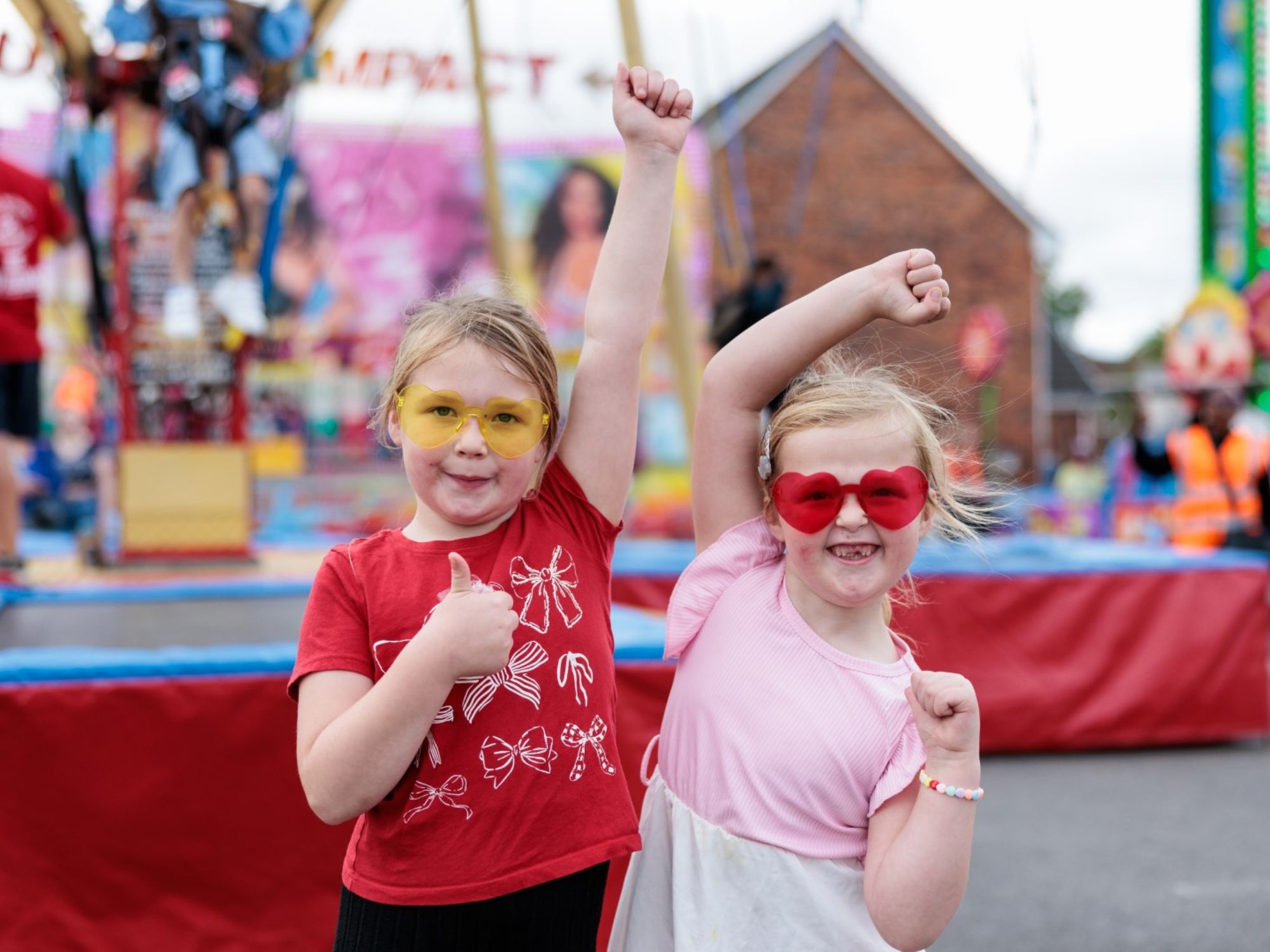 Two children posing joyfully at a colorful fairground. One wears a red shirt with white bows and yellow-tinted glasses, giving a thumbs-up. The other wears a pink shirt and red heart-shaped glasses, raising both arms in celebration. Blurred amusement rides and decorations fill the background.