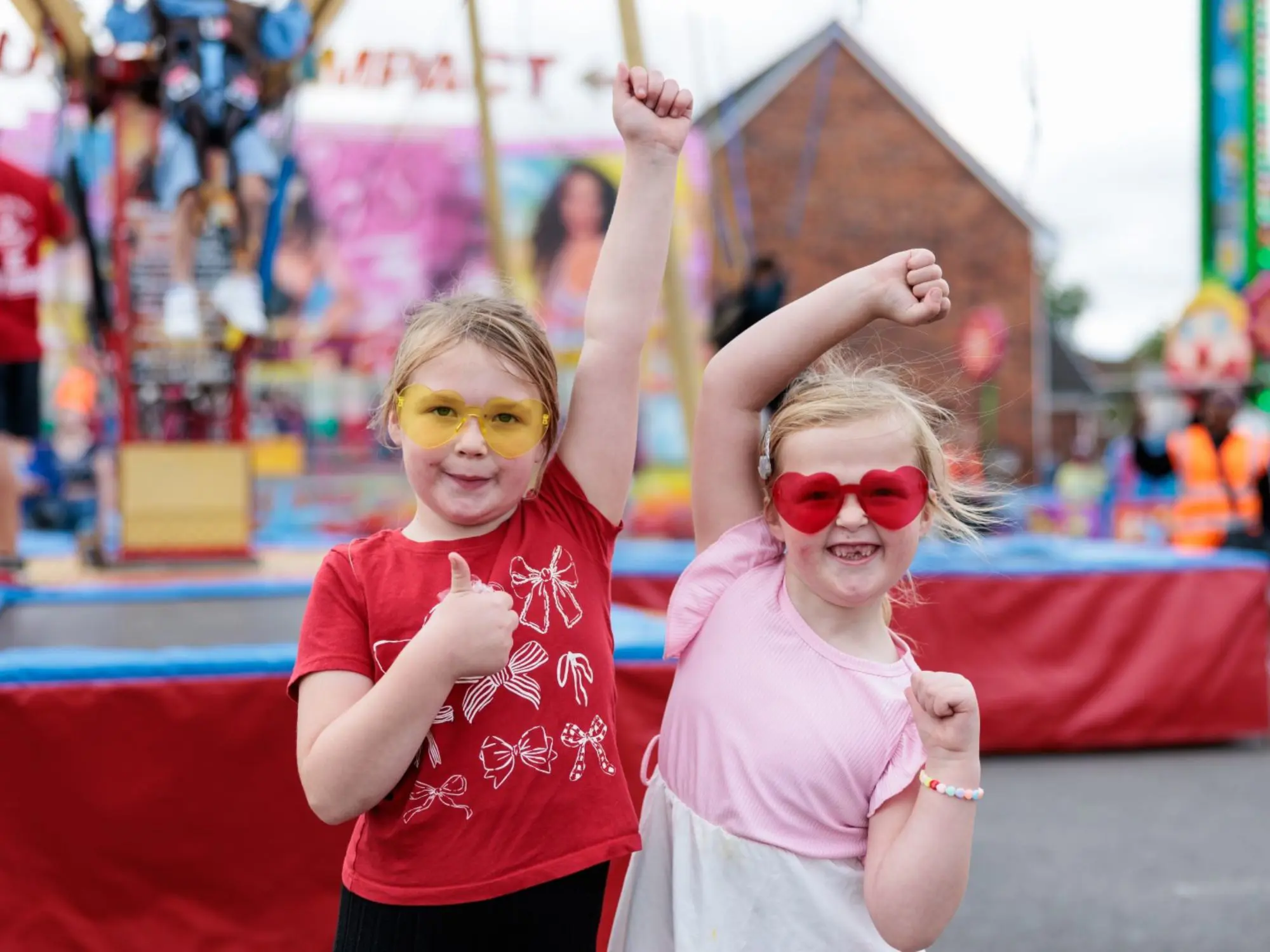 Two children posing joyfully at a colorful fairground. One wears a red shirt with white bows and yellow-tinted glasses, giving a thumbs-up. The other wears a pink shirt and red heart-shaped glasses, raising both arms in celebration. Blurred amusement rides and decorations fill the background.