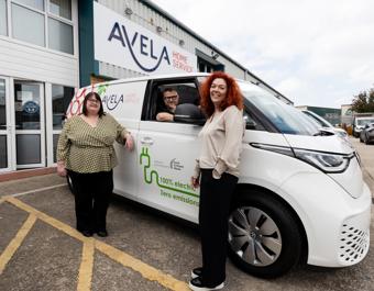 Three people standing beside a white AVELA Home Service electric van with '100% electric, zero emissions' written on the side. The AVELA Home Service building and company signage are visible in the background.
