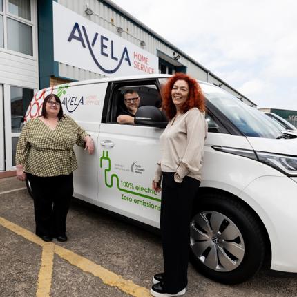 Three people standing beside a white AVELA Home Service electric van with '100% electric, zero emissions' written on the side. The AVELA Home Service building and company signage are visible in the background.