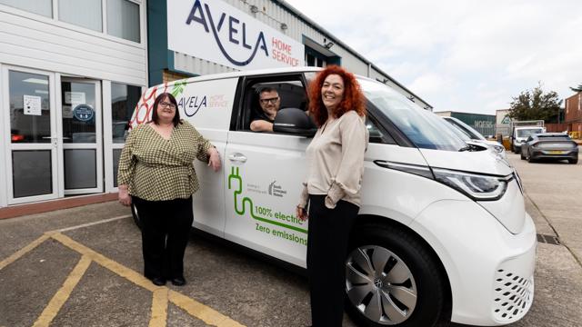 Three people standing beside a white AVELA Home Service electric van with '100% electric, zero emissions' written on the side. The AVELA Home Service building and company signage are visible in the background.