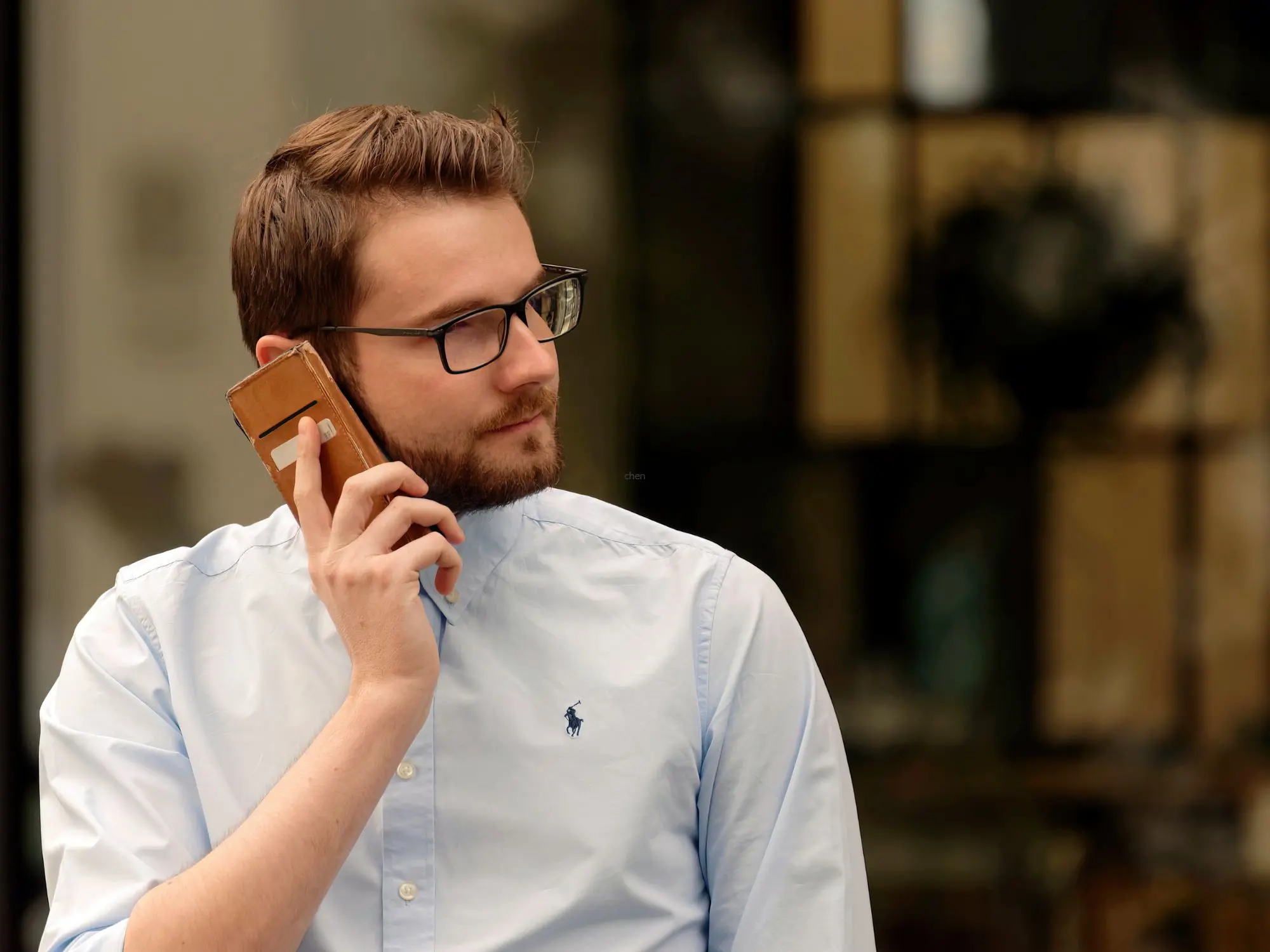 Person wearing glasses and a light blue shirt talking on a smartphone outdoors, with reflections of trees and buildings in the background.