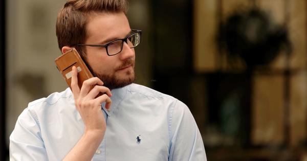 Person wearing glasses and a light blue shirt talking on a smartphone outdoors, with reflections of trees and buildings in the background.