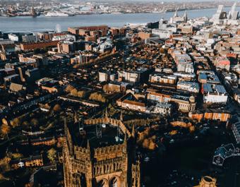 Aerial view of Liverpool cityscape with a prominent cathedral in the foreground, surrounded by dense urban buildings, roads, green spaces, and the River Mersey with industrial structures and ships in the background.