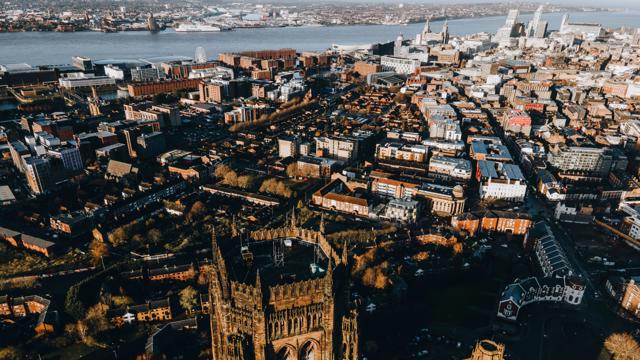 Aerial view of Liverpool cityscape with a prominent cathedral in the foreground, surrounded by dense urban buildings, roads, green spaces, and the River Mersey with industrial structures and ships in the background.