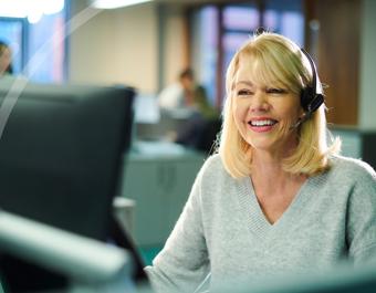 A person with blonde hair wearing a headset sits at a desk in an office environment. Other people and office equipment are visible in the background.