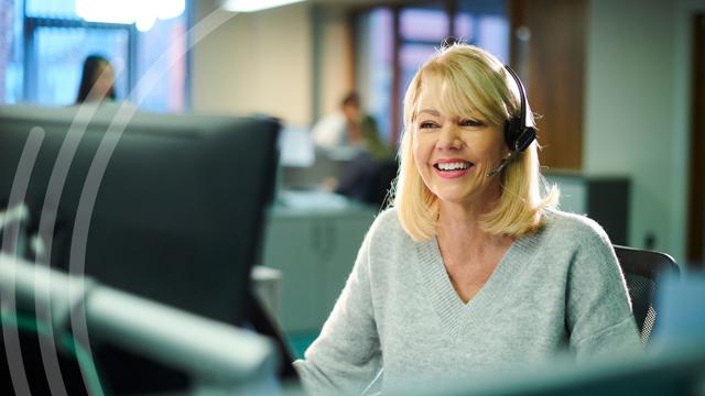 A person with blonde hair wearing a headset sits at a desk in an office environment. Other people and office equipment are visible in the background.