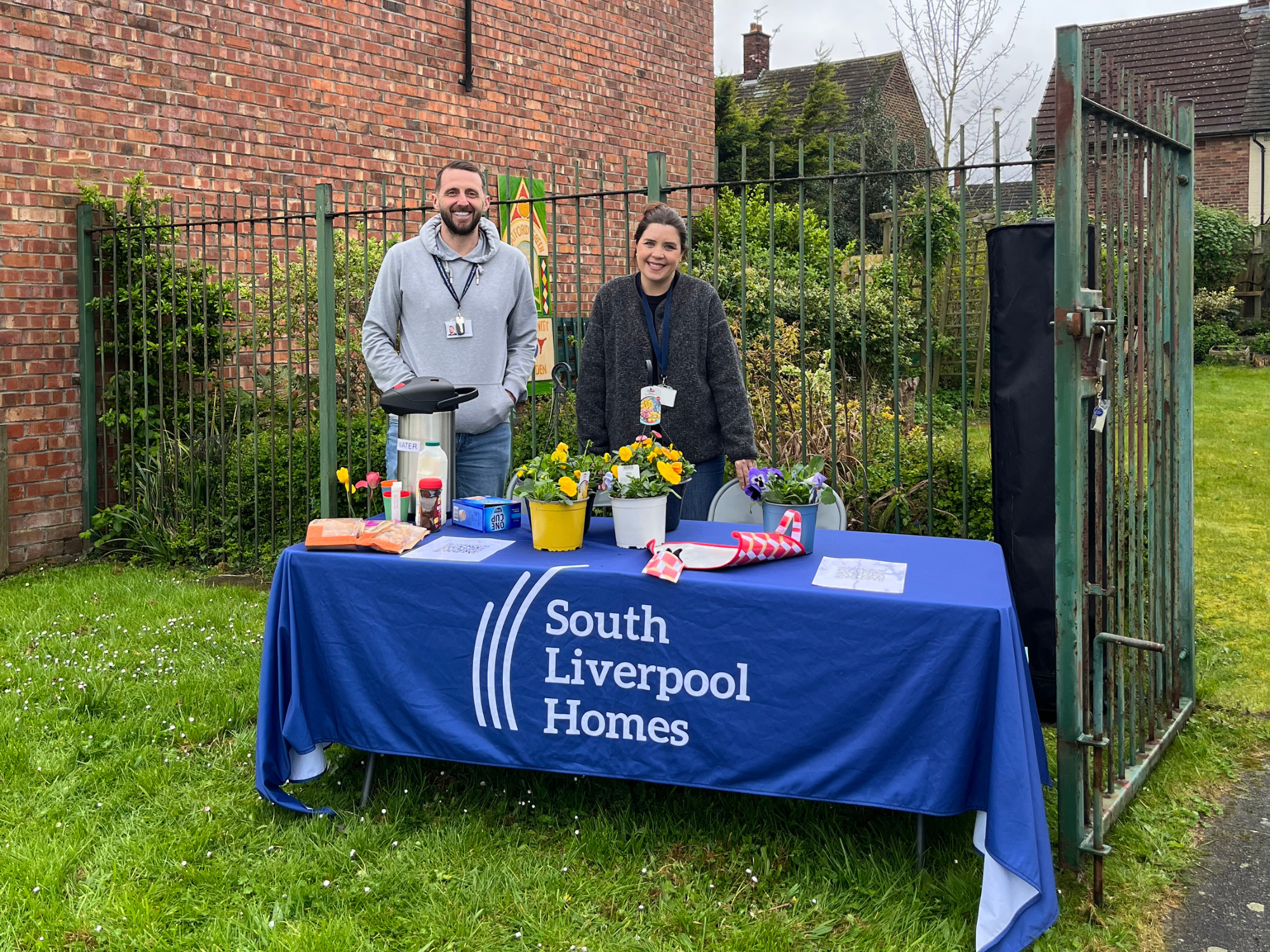 Two people standing behind a table covered with a South Liverpool Homes tablecloth at an outdoor community event. The table displays potted flowers, gardening items and refreshments. A fenced garden and brick building are visible in the background.