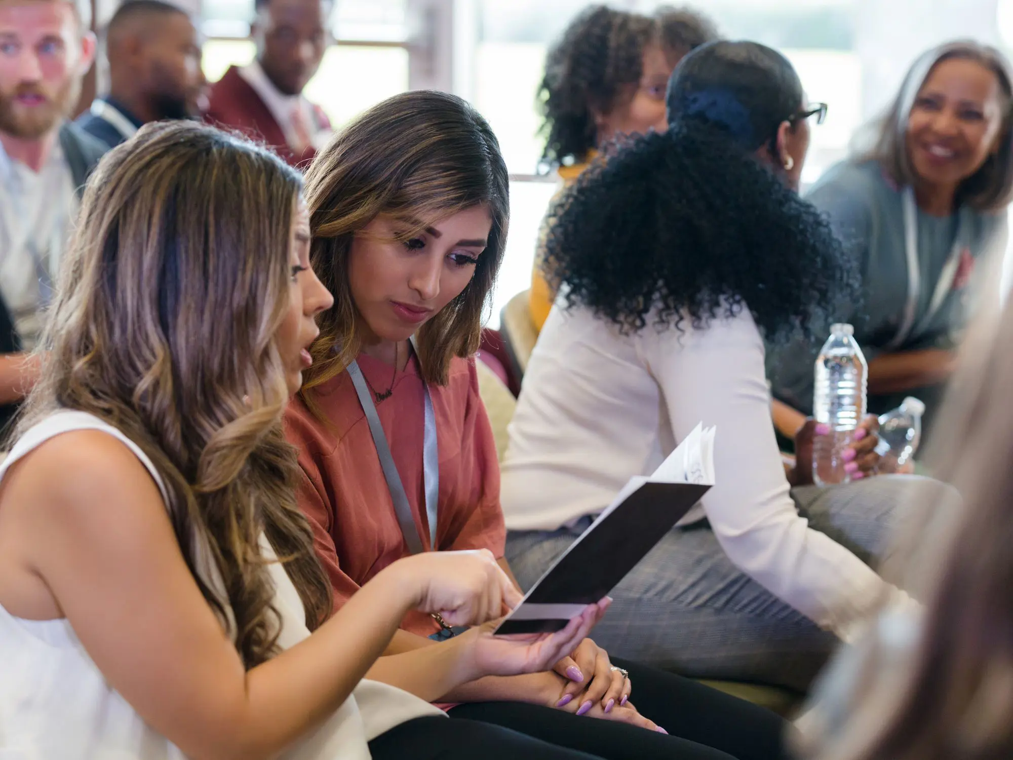 People seated in a conference setting, with two individuals in the foreground discussing a document or tablet, surrounded by others engaged in conversation, reflecting a collaborative and interactive environment.