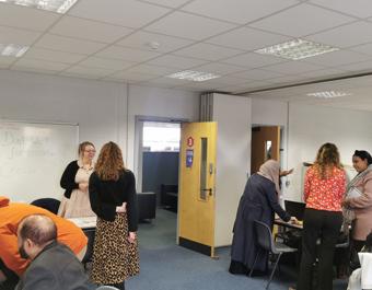 Group of people interacting in a classroom or office setting with tables, chairs, and a whiteboard displaying the message: 'Don't wait for opportunity. Create it!