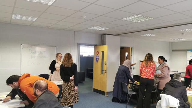 Group of people interacting in a classroom or office setting with tables, chairs, and a whiteboard displaying the message: 'Don't wait for opportunity. Create it!
