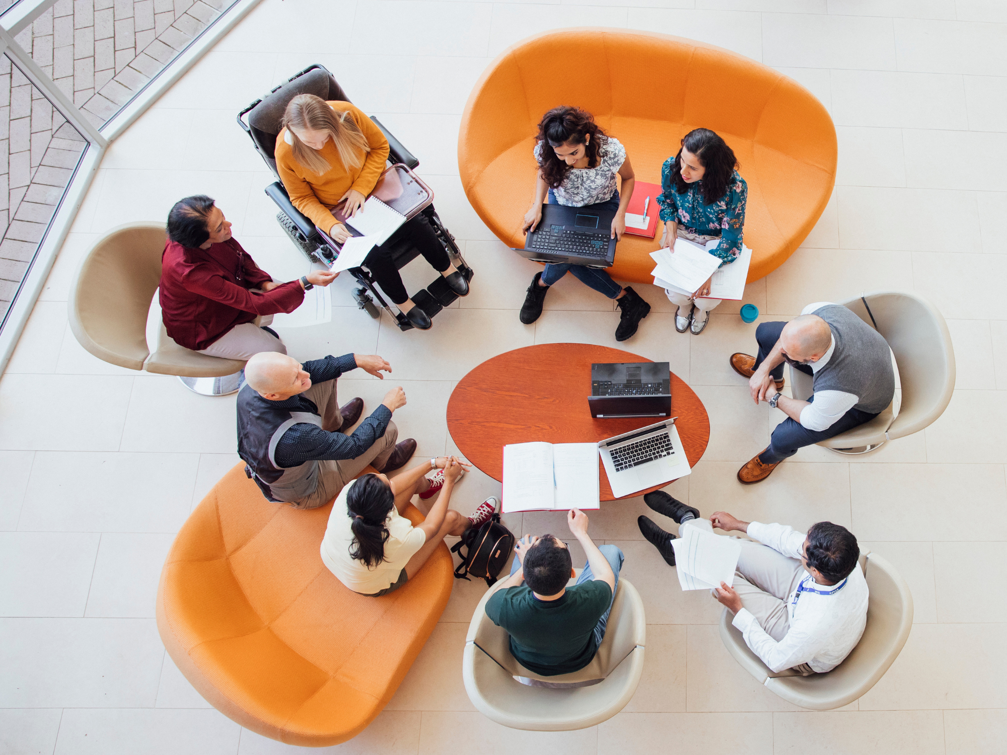 Overhead view of a group seated in a circle on sofas and chairs, discussing documents and laptops around a small round table, including one wheelchair user.
