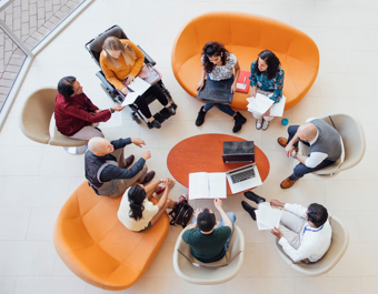 Overhead view of a group seated in a circle on sofas and chairs, discussing documents and laptops around a small round table, including one wheelchair user.