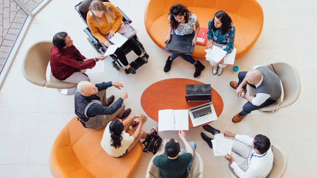 Overhead view of a group seated in a circle on sofas and chairs, discussing documents and laptops around a small round table, including one wheelchair user.