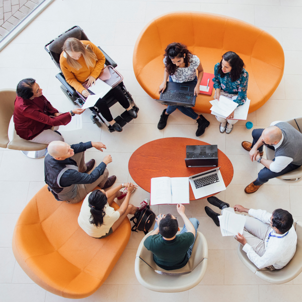 Overhead view of a group seated in a circle on sofas and chairs, discussing documents and laptops around a small round table, including one wheelchair user.