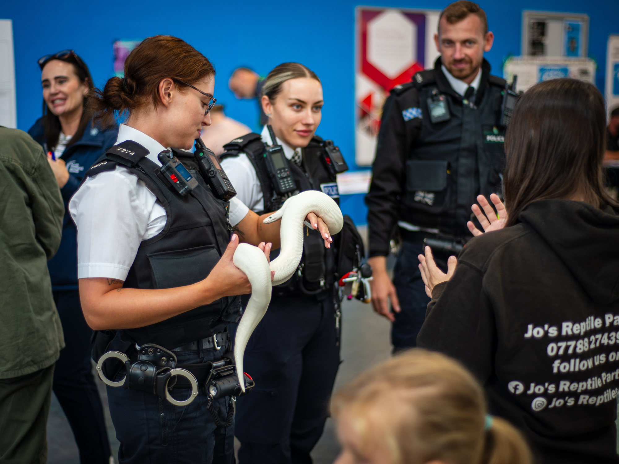 Police officers in uniform speak with members of the public at an indoor event, as one officer holds a white snake and shows it to a small group. A person wearing a black hoodie with “Jo’s Reptile Parties” on the back stands nearby, while other attendees are visible in the background.
